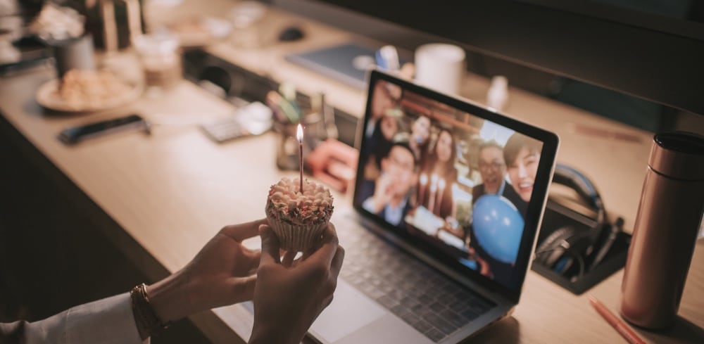 person holding a cupcake while colleages on a computer screen are celebrating with balloons