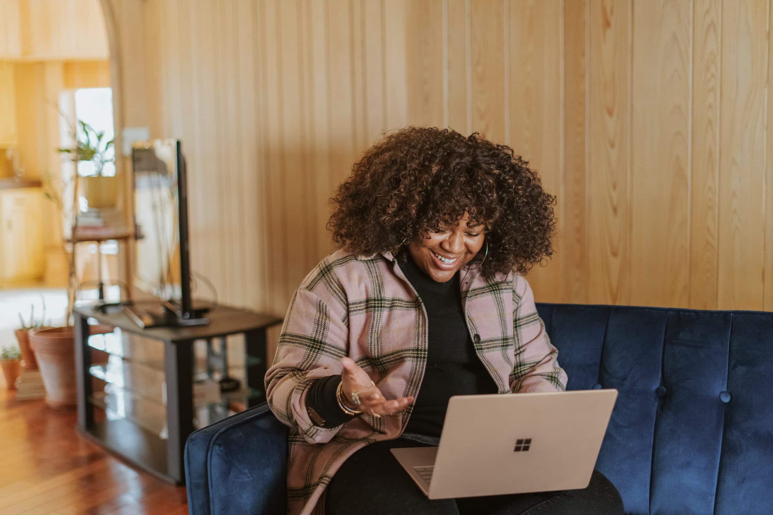 Black woman smiling on a blue couch engaging in a video call on her laptop