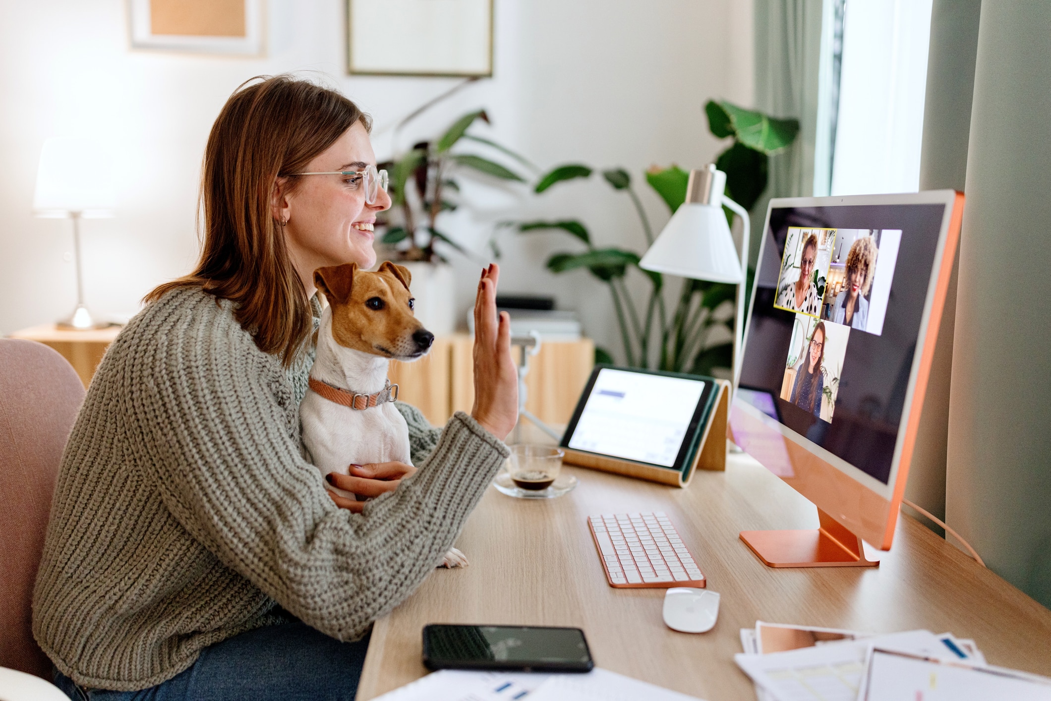 Woman waving at coworkers on a video call meeting at home. She is sitting at her desk with a small brown and white dog in her lap.