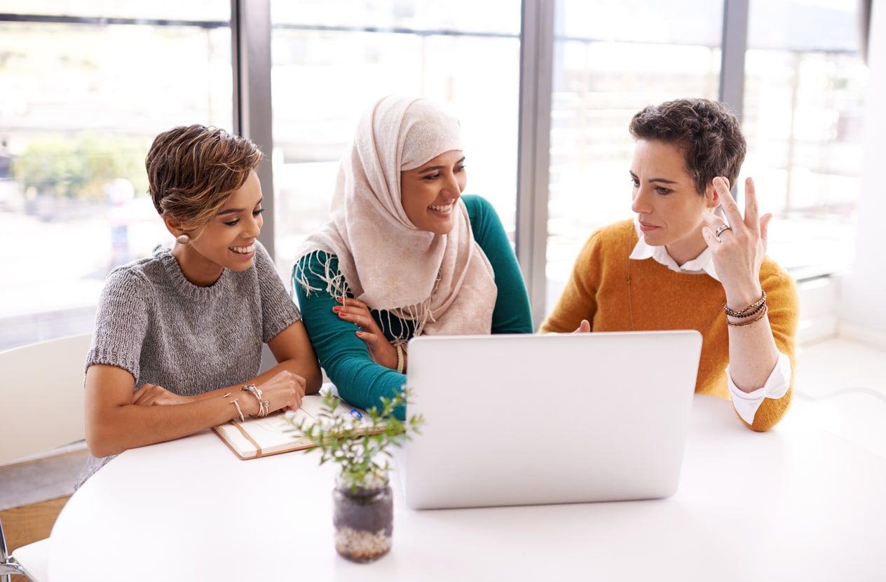 Group of diverse women at an office meeting, smiling and talking in front of a laptop.